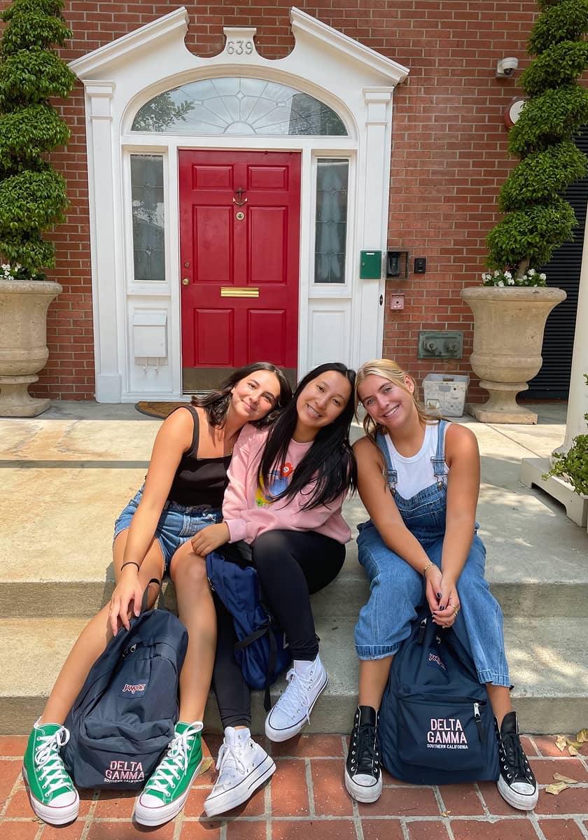 Three girls sitting for a photo in front of red door with backpacks