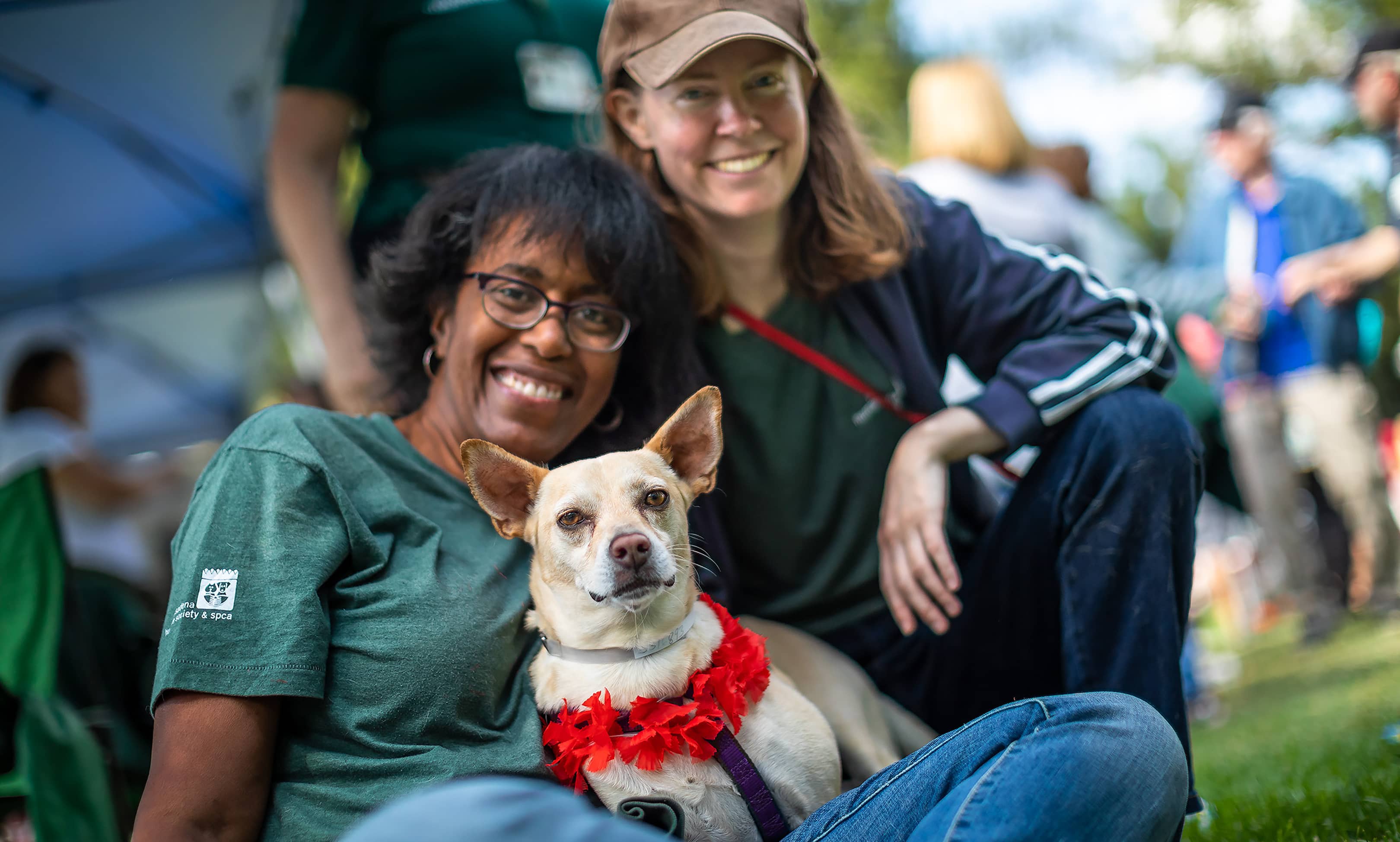Two women sitting on the ground with a chihuahua in their lap smiling at the camera.