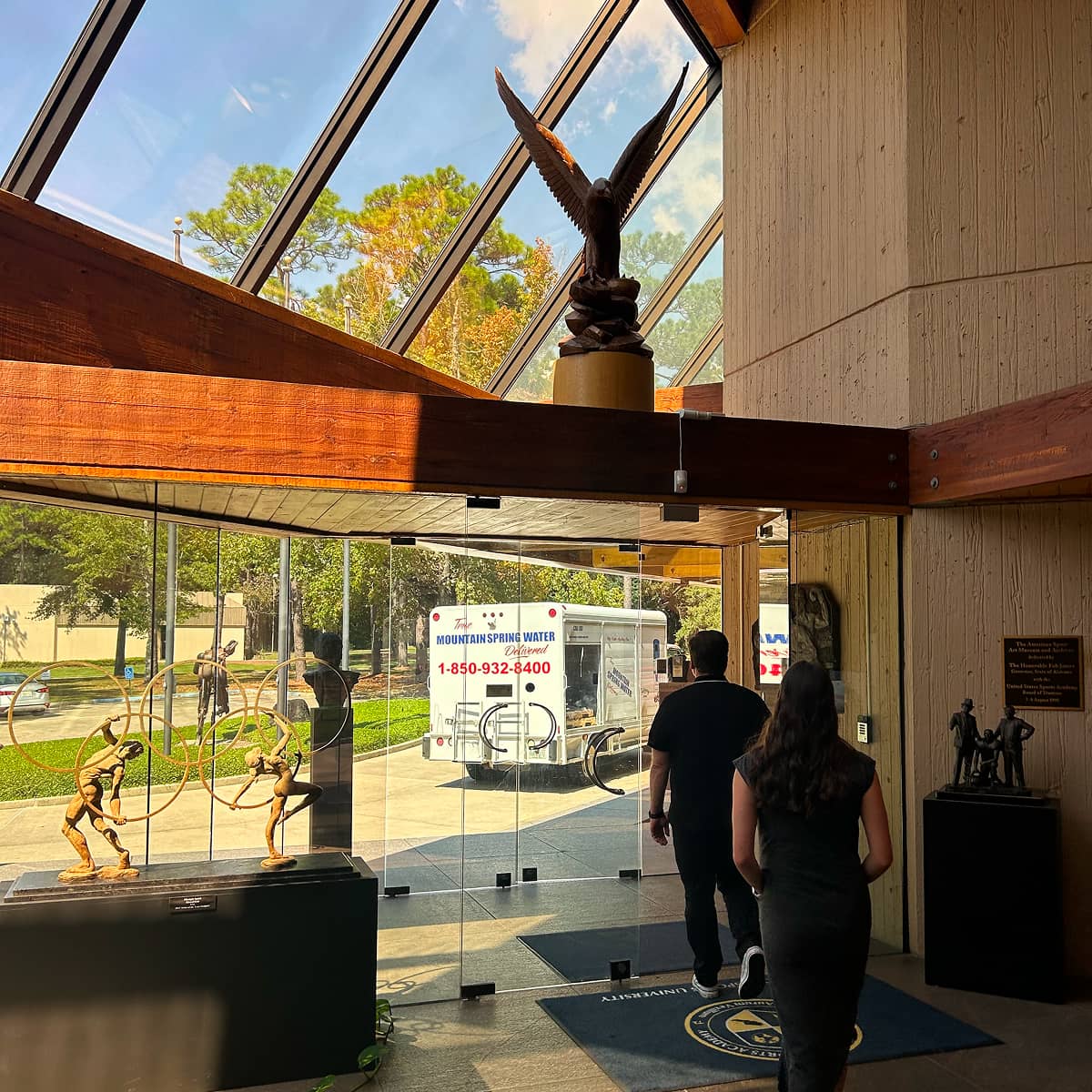 Indoor scene with two people walking toward a glass door, sunlight streaming through large angled skylights. Sculptures are visible on both sides.