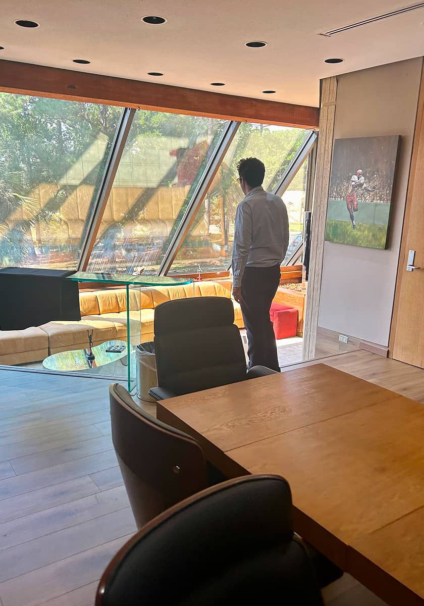 A person in business attire stands by slanted glass windows, overlooking trees. The room has modern furniture and a sports-themed painting on the wall.