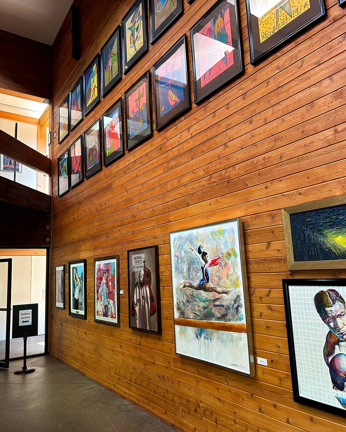 Wood-paneled gallery wall with colorful paintings and framed art. The room feels inviting and vibrant, showcasing diverse artistic styles and subjects.