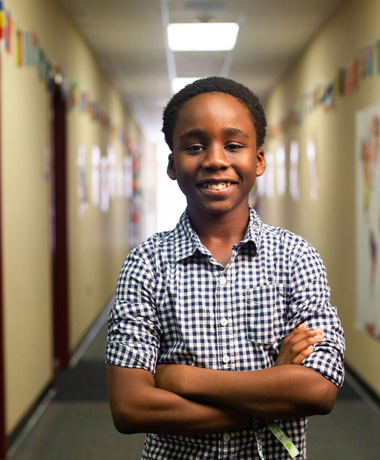 A young boy in a checkered shirt stands confidently with crossed arms in a school hallway. The bright lighting creates a welcoming atmosphere.