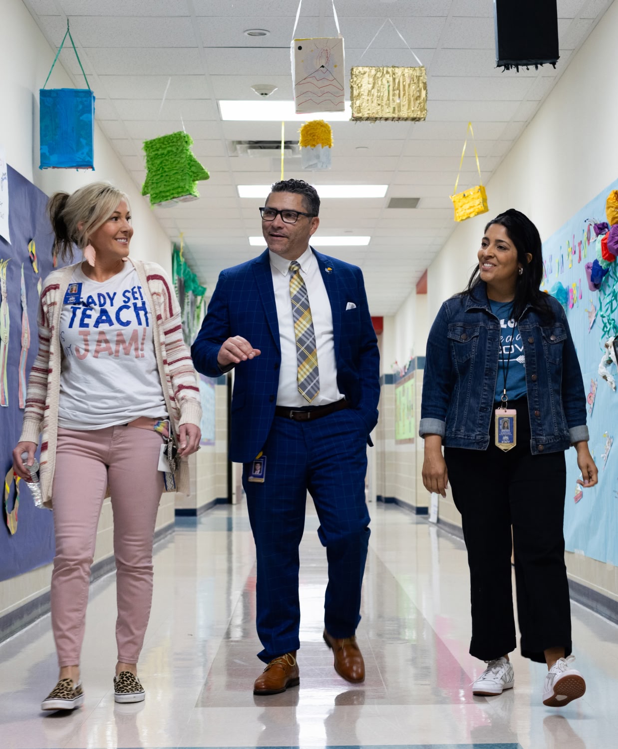 Three people walk down a decorated school hallway, smiling and engaged in conversation. The atmosphere is cheerful and collaborative.