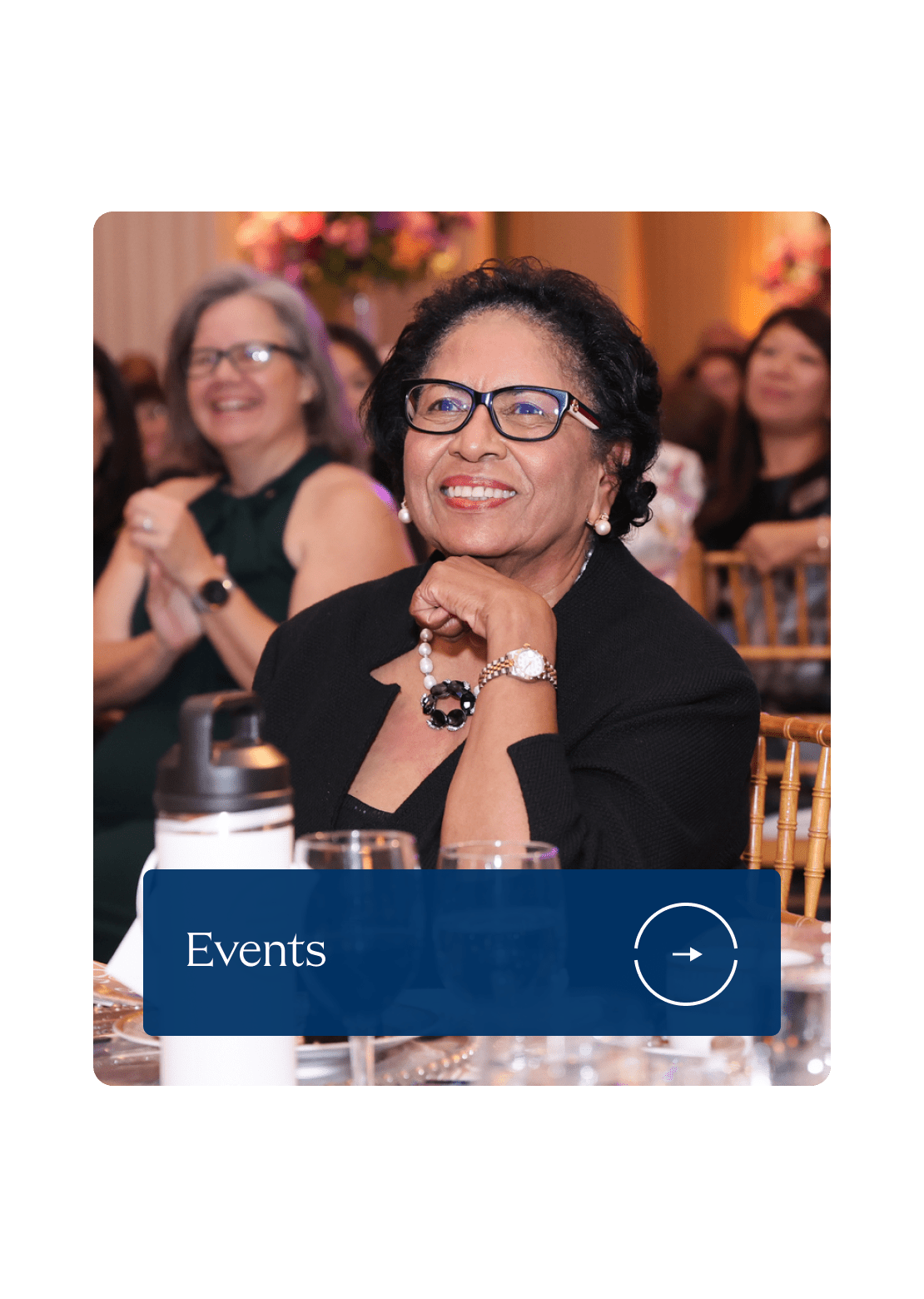 A smiling woman in glasses sits at a formal event, surrounded by applauding attendees. The atmosphere is joyful. Text overlay reads 