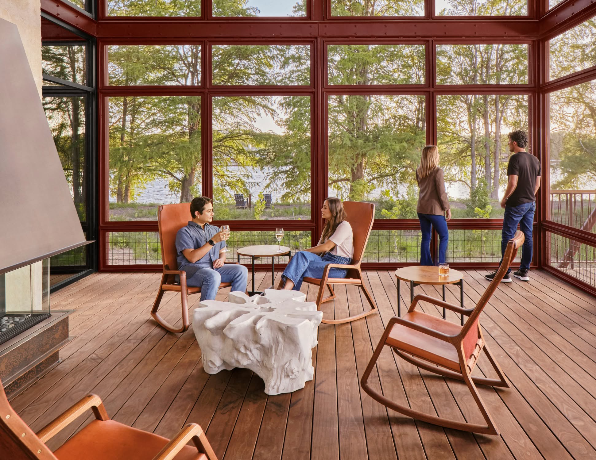 A cozy room with large windows offers a scenic lake view. Two people sit chatting with drinks, while two others enjoy the view, creating a relaxed atmosphere.