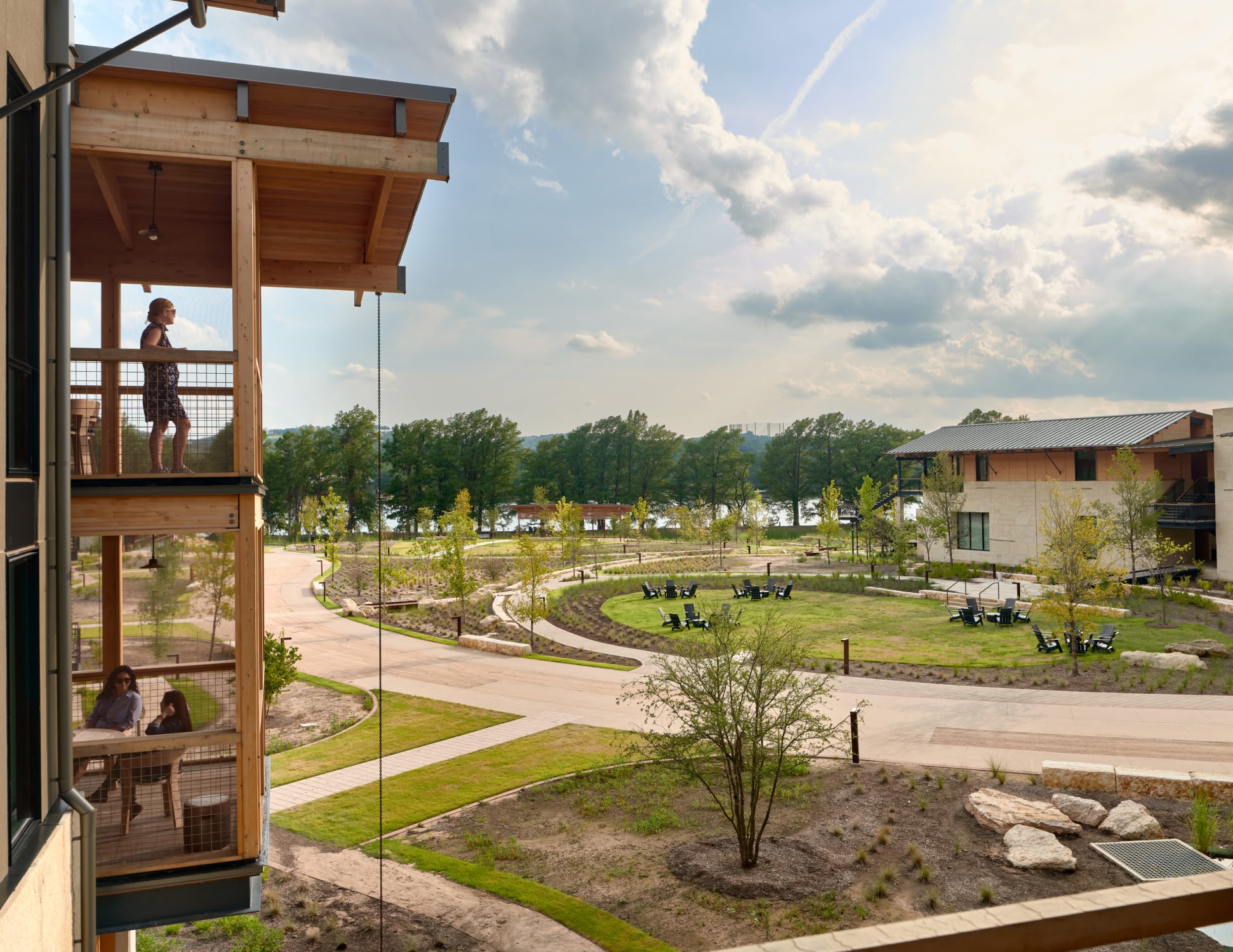 Wooden balconies overlook a sunlit park with scattered trees and outdoor seating. A peaceful, expansive landscape under a partly cloudy sky.