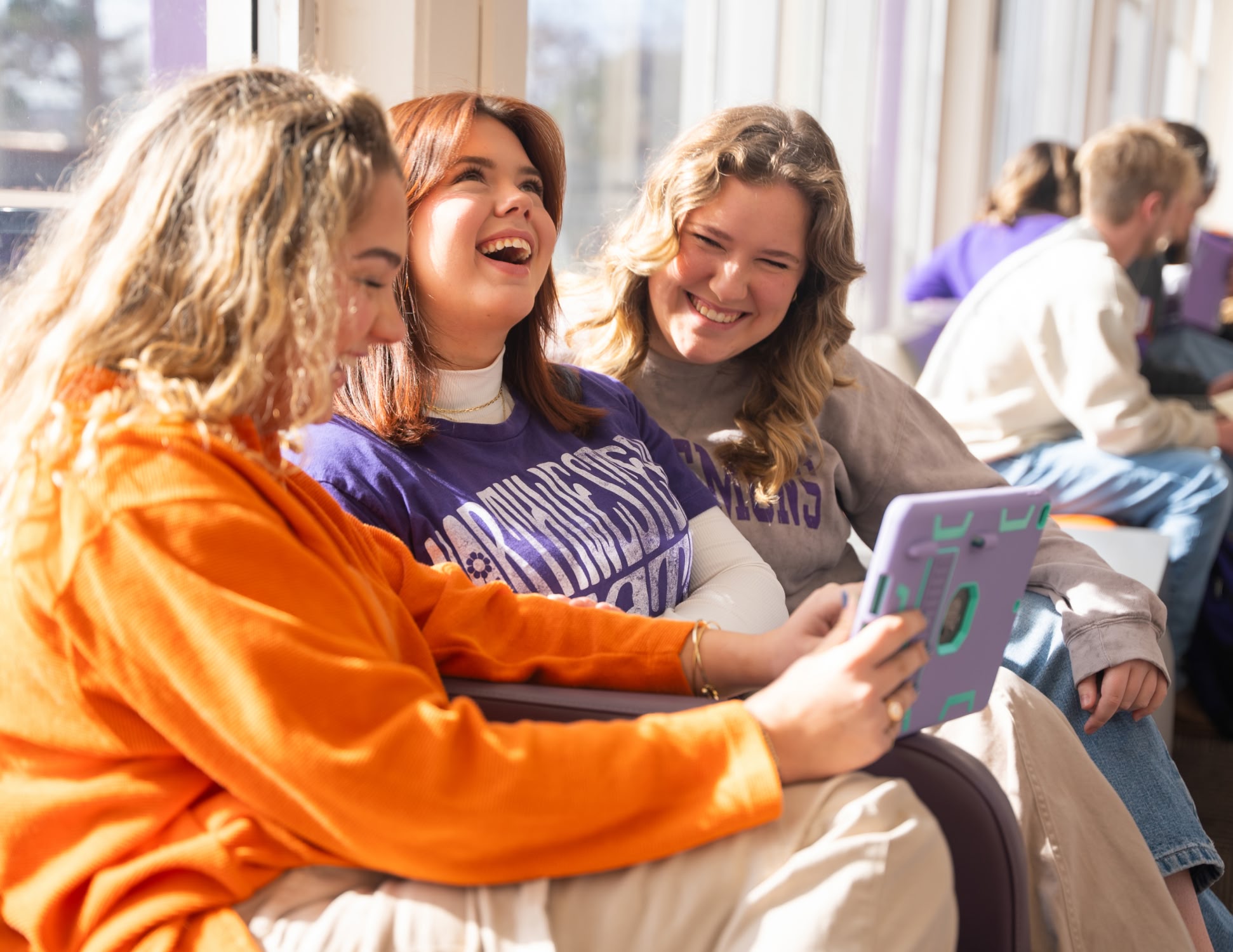 Three young women sit together, laughing and looking at a tablet. They wear colorful casual clothing in a brightly lit room, creating a joyful and lively atmosphere.