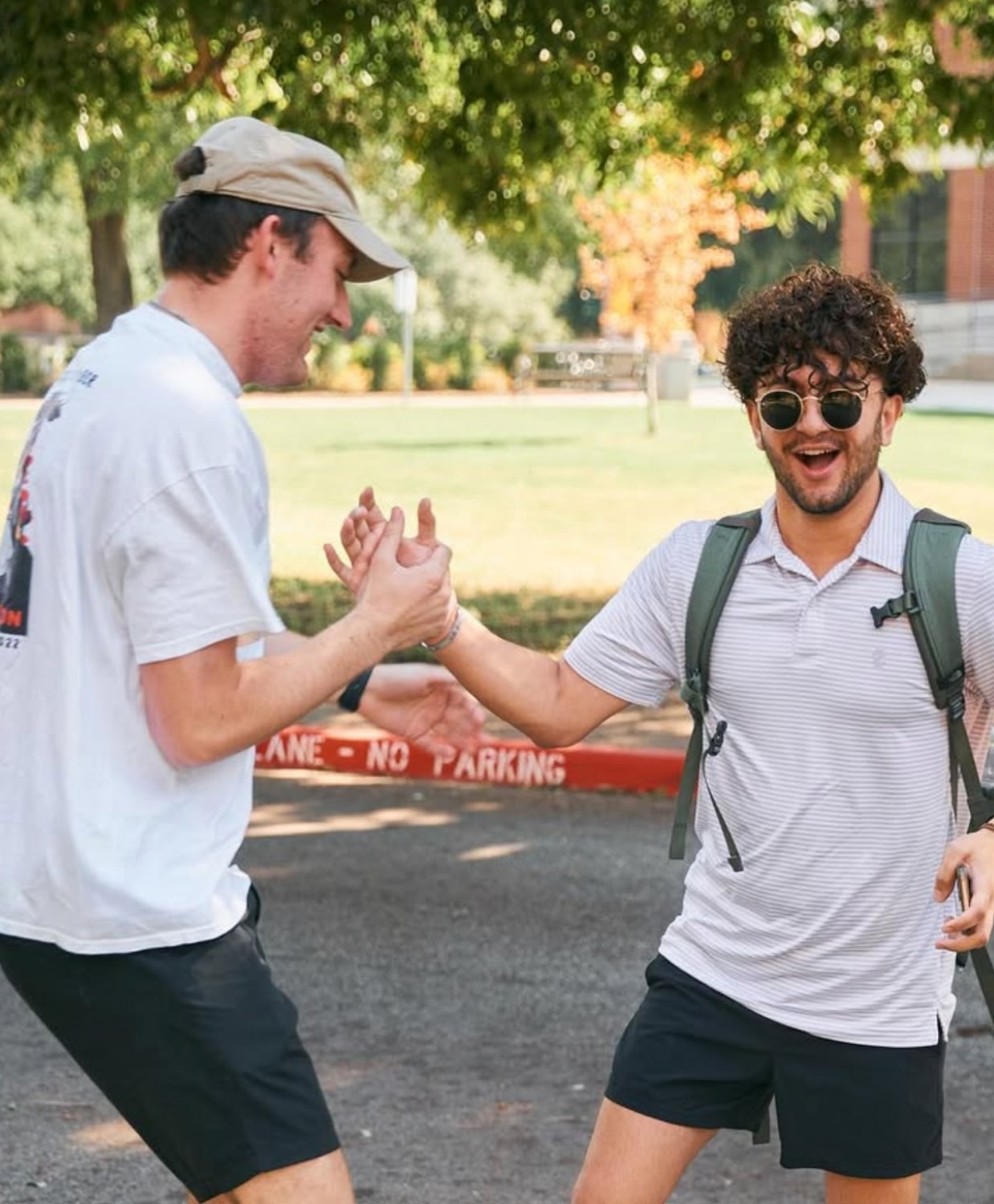 Two young men smiling, engaging in a friendly handshake outdoors. They wear casual summer attire, with trees and a 
