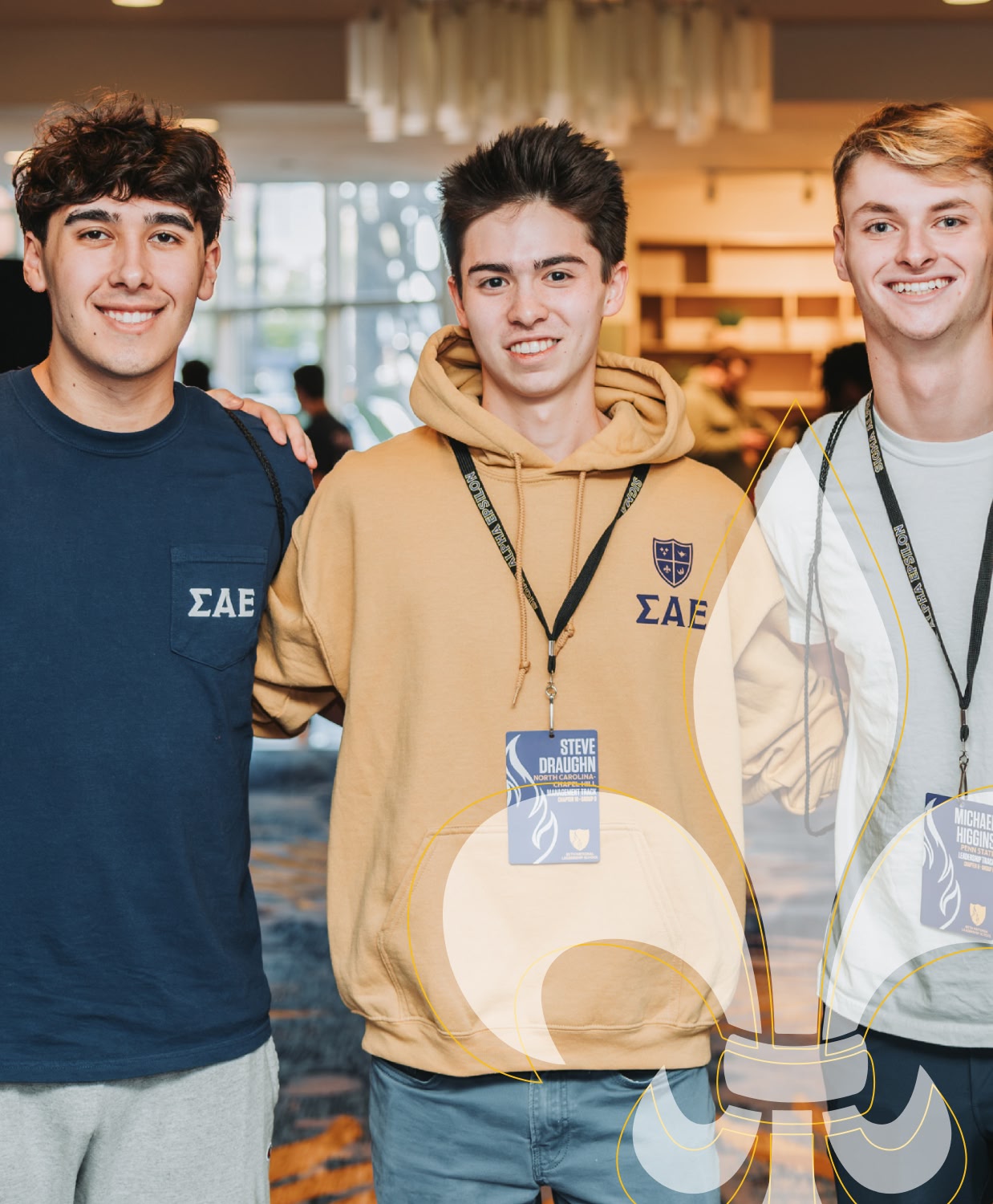 Three young men smiling and posing indoors. They wear lanyards and clothing with Greek letters. The mood is friendly and upbeat.