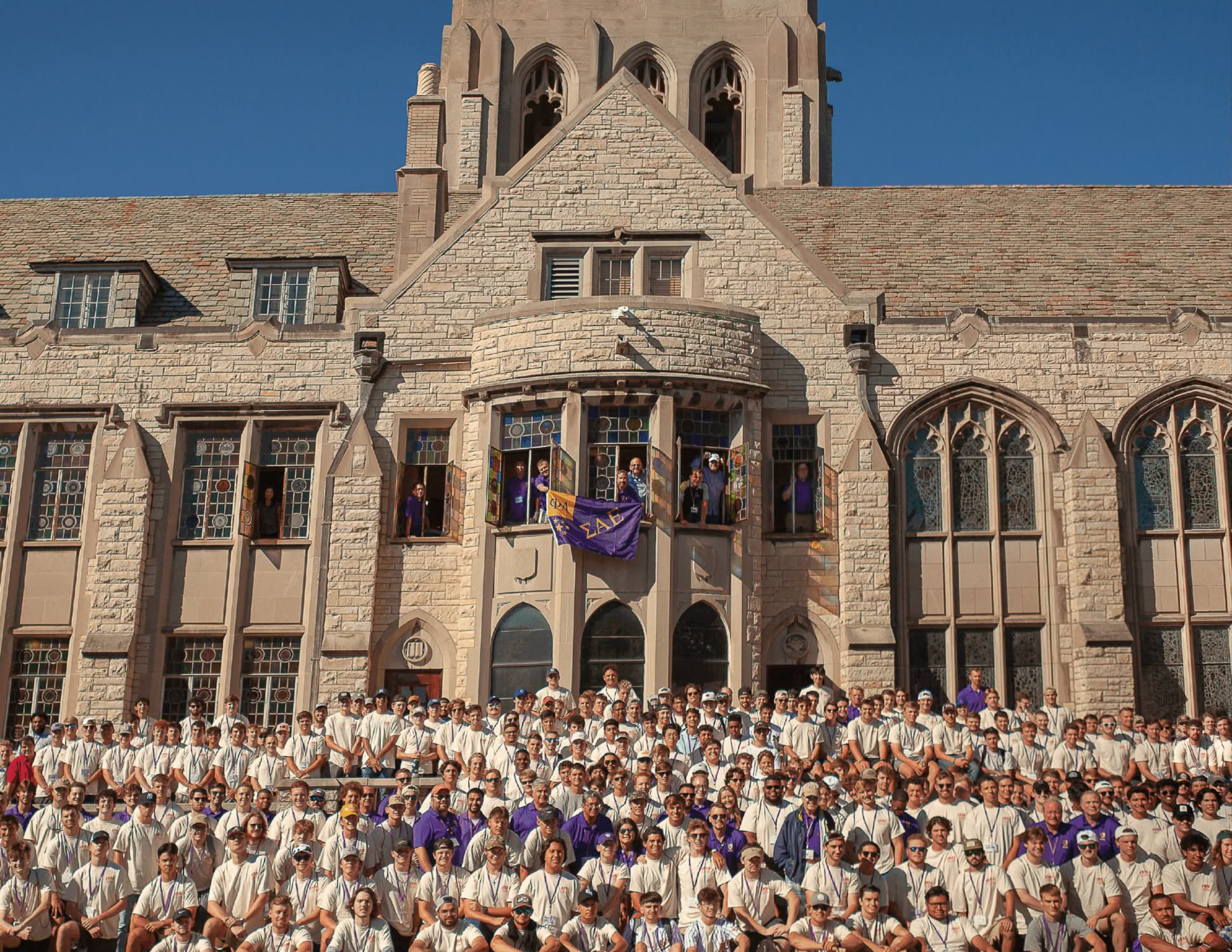 A large group of people in matching outfits pose for a photo in front of a historic stone building with tall arched windows. A purple flag hangs above.
