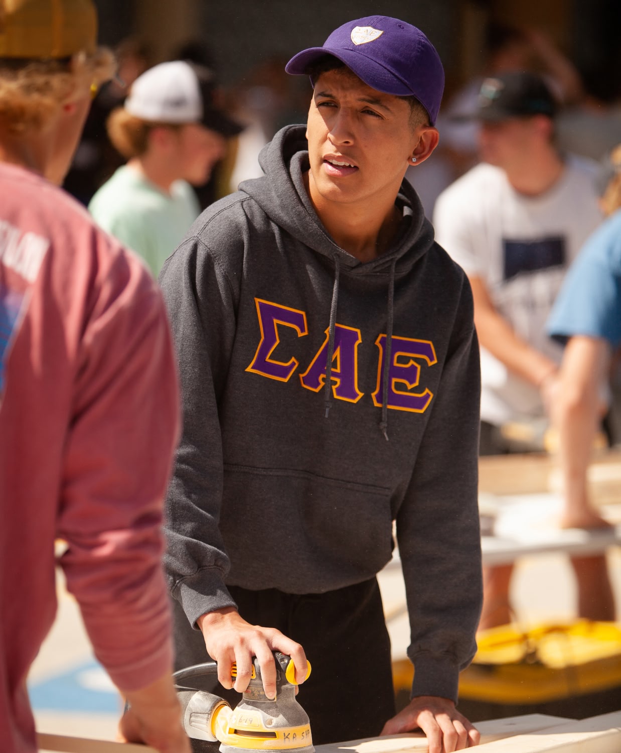 A young man wearing a purple cap and a gray Sigma Alpha Epsilon hoodie sands wood outdoors, looking focused. Others work in the blurred background.