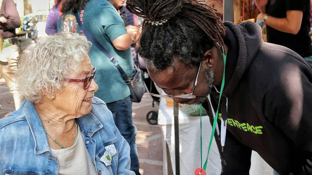 An older woman and a nonprofit volunteer share a conversation at an outdoor community event, reflecting the vibrant mission-driven work aging services organizations deliver every day.