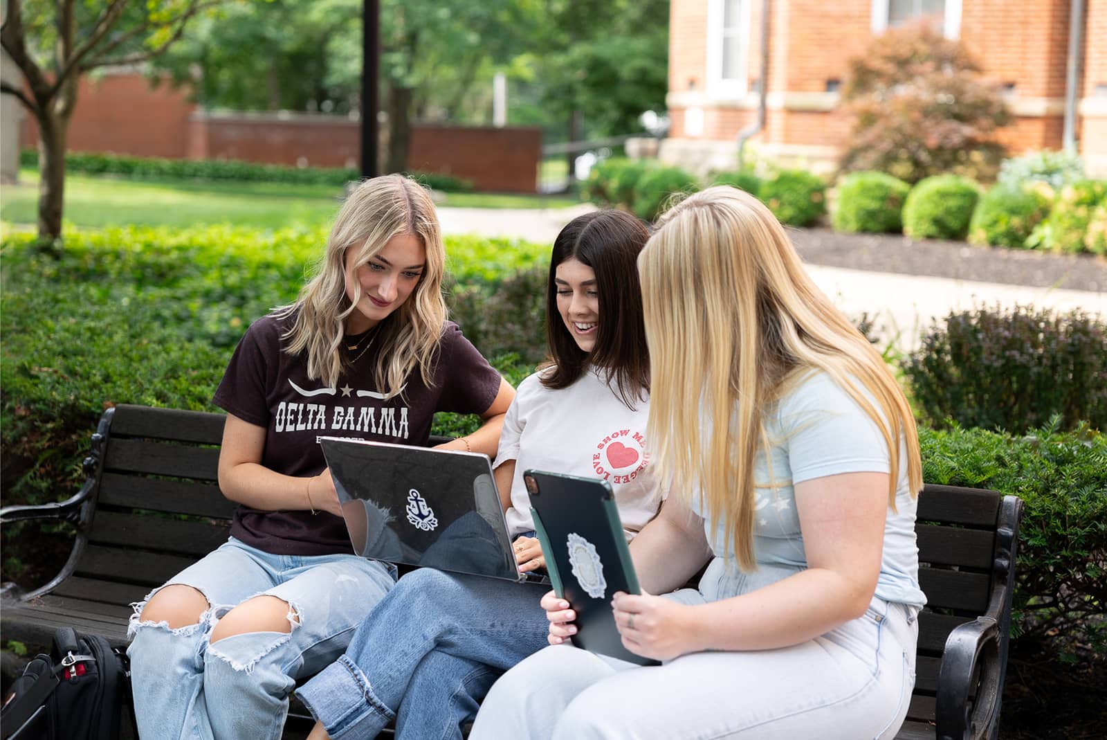 Three Delta Gamma members sit together on a campus bench, laughing and sharing content on a laptop and tablet, wearing Delta Gamma branded apparel.