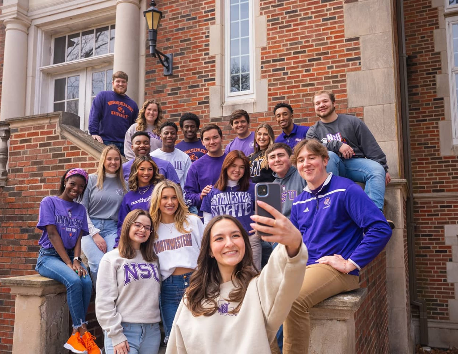 A diverse group of smiling college students wearing Northwestern State University apparel gather on brick steps for a selfie, exuding a cheerful, collegiate vibe.