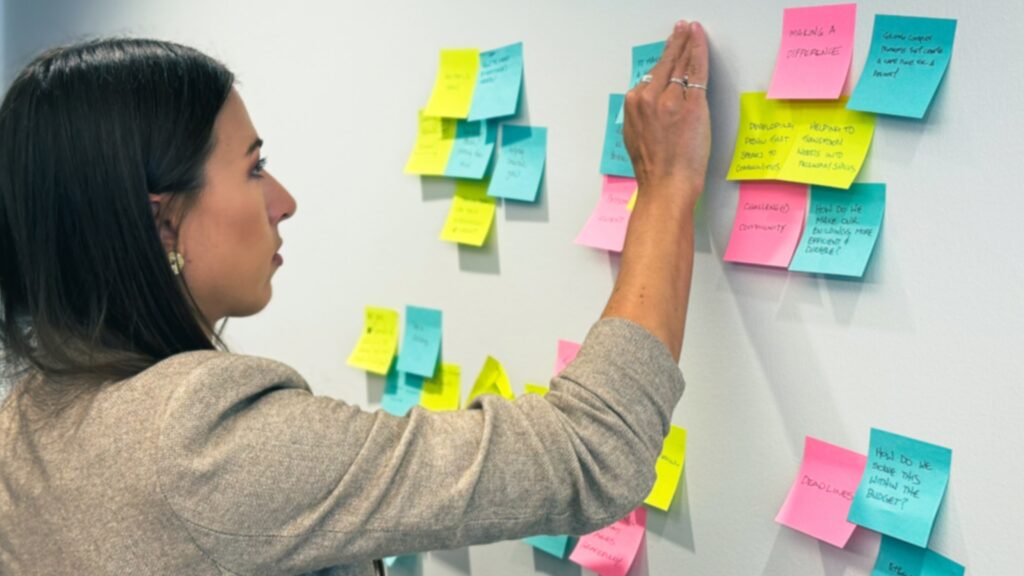 Sabrina arranges colorful sticky notes on a wall, focusing intently. Notes are pink, blue, and yellow, indicating brainstorming or planning.