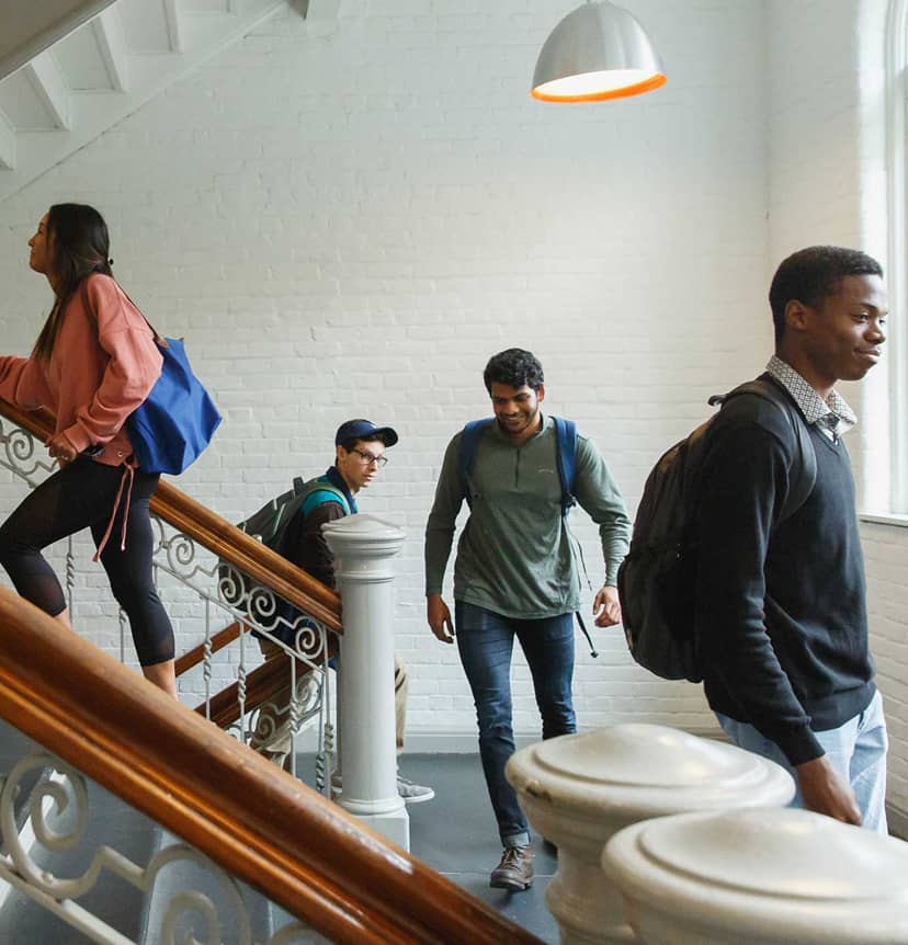 Students walking down a bright staircase with white brick walls and wood banisters, backpacks in hand, heading to class.