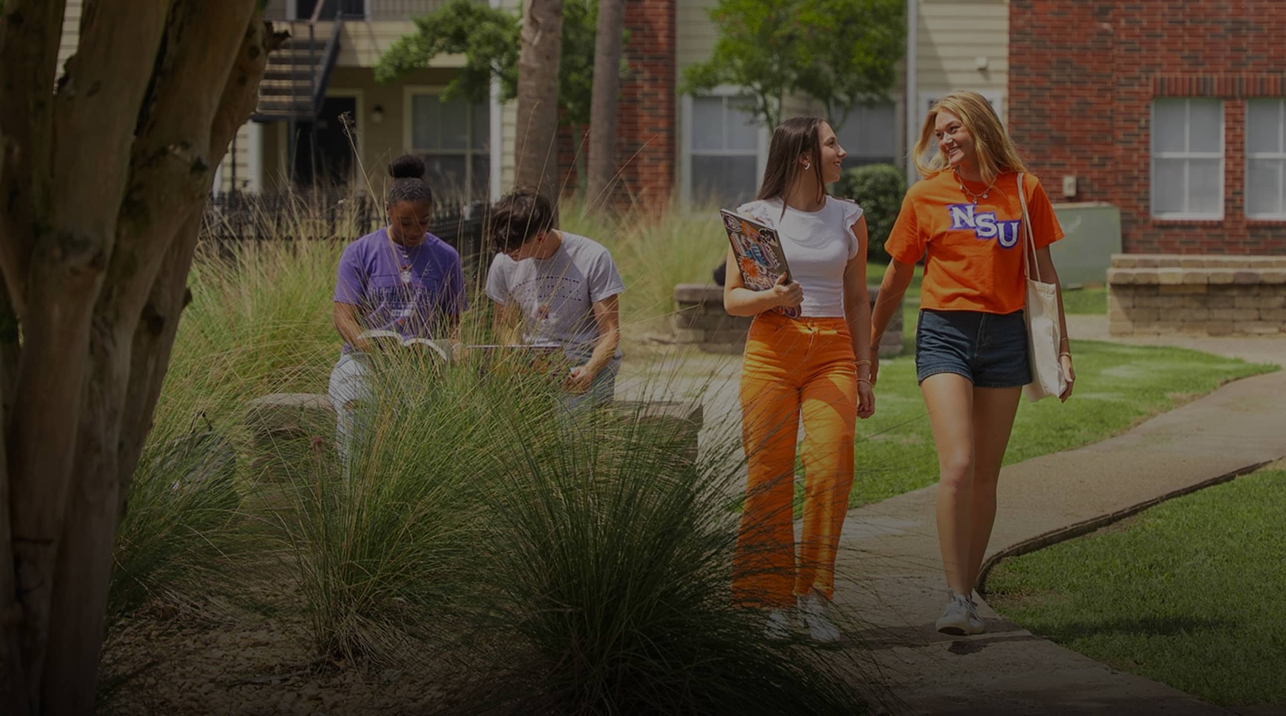 Northwestern State University students walking and talking on a campus sidewalk, with two others studying on a bench in the background.