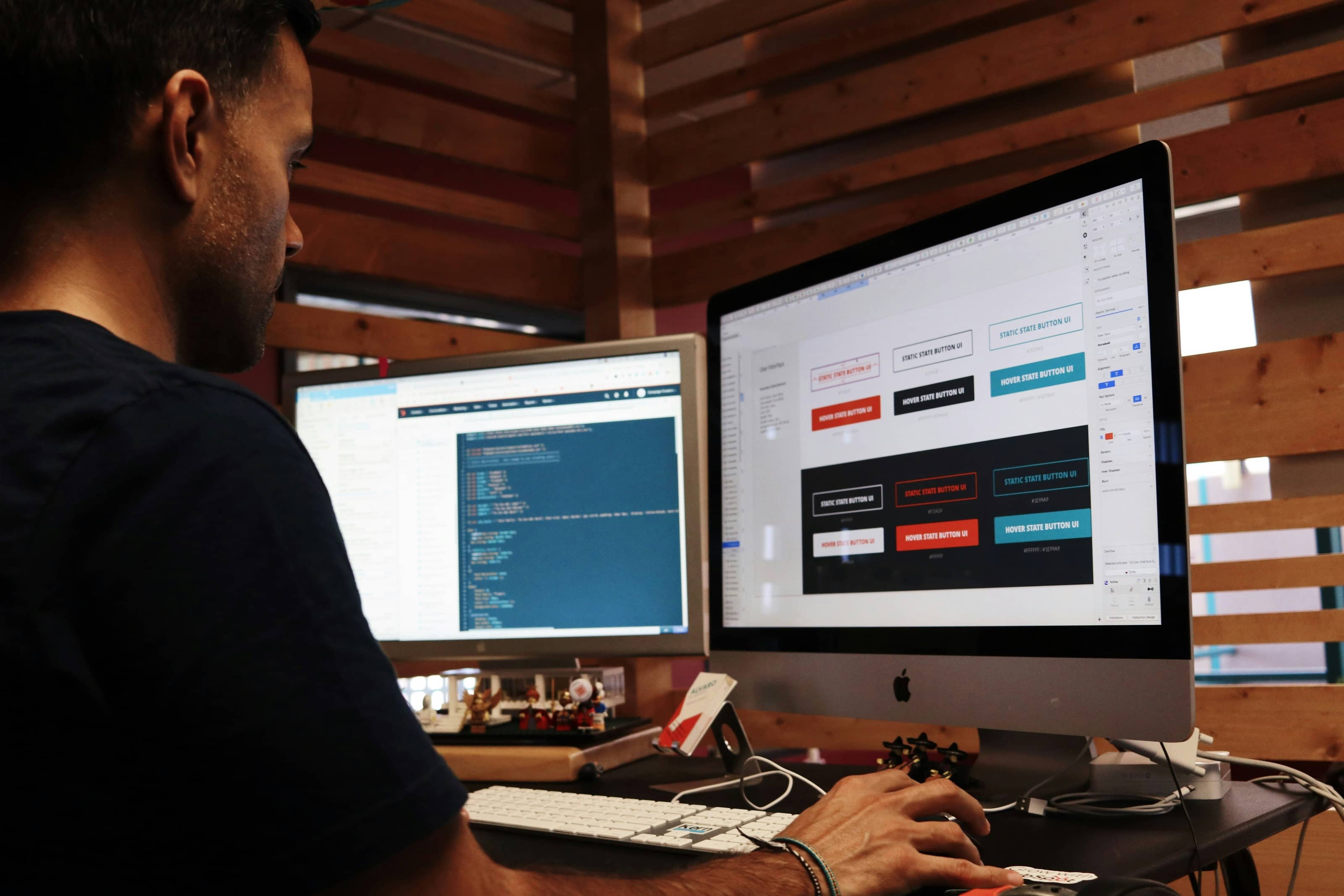 Developer working at a dual-monitor desk, with code displayed on one screen and UI button design states on the other.
